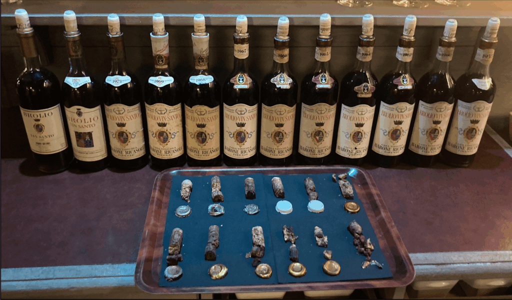 Une verticale de Vin Santo du Castello di Brolio, millésimes 1980 à 1955, alignée sur un comptoir, accompagnée d’un plateau présentant les bouchons et capsules de chaque bouteille, dans un cadre sobre et élégant.