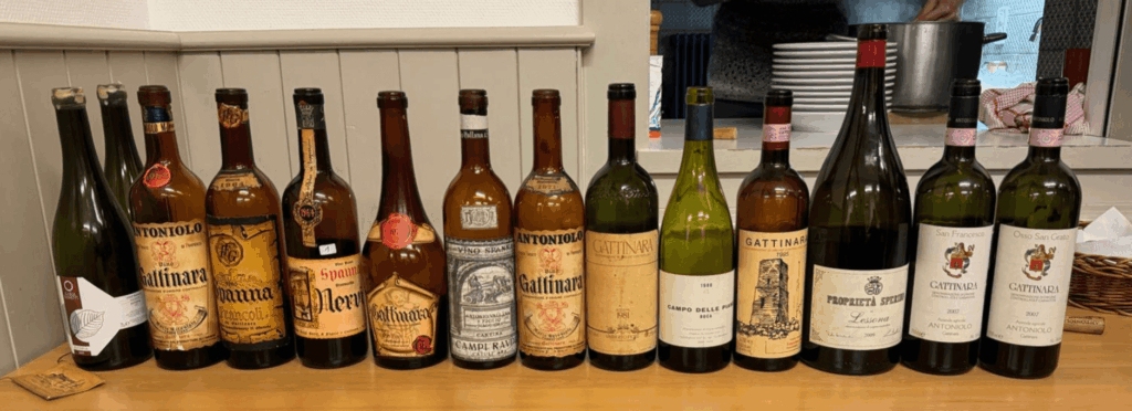 Une sélection de vins du Haut Piémont, incluant des Gattinara, Boca et Lessona des millésimes 1962 à 2009, alignés sur un comptoir en bois, avec étiquettes vieillies et bouchons colorés, devant un mur blanc à panneaux verticaux.