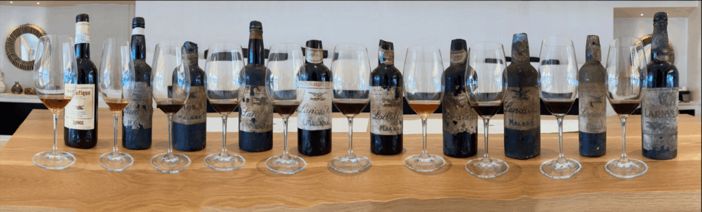 Une série de bouteilles de Malaga anciennes, millésimes allant jusqu’en 1817, alignées sur un comptoir en bois avec des verres remplis de vin, devant un mur blanc et un miroir décoratif.