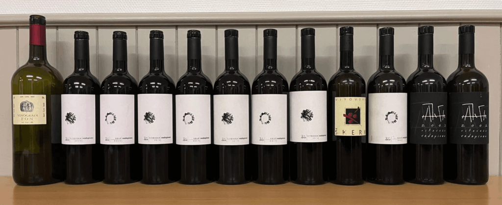 Une série de bouteilles de Vitovska du Carso, millésimes 2002 à 2020, incluant les cuvées de Paolo Vodopivec et Sandi Skerk, alignées sur un comptoir en bois, avec étiquettes minimalistes et capsules noires, devant un mur blanc à panneaux verticaux.