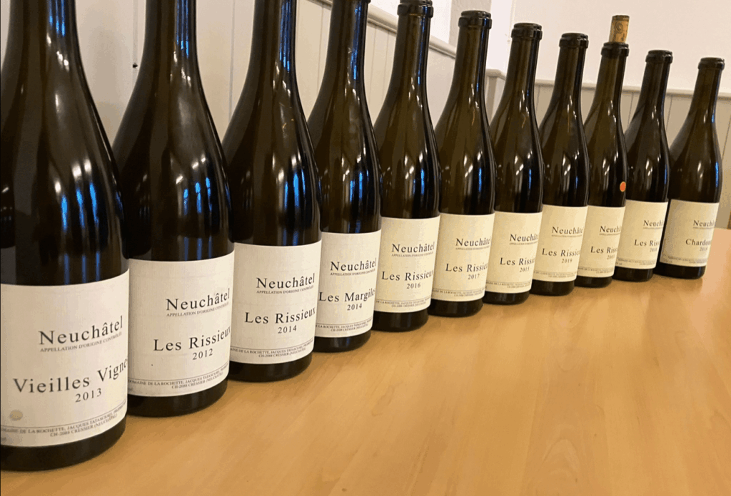 Une verticale de vins Neuchâtel du Domaine de la Rochette, incluant les cuvées Les Rissieux, Les Margiles et Chardonnay, millésimes 2012 à 2019, alignés sur une table en bois clair, avec étiquettes minimalistes et bouchons noirs, devant un mur blanc à panneaux verticaux.