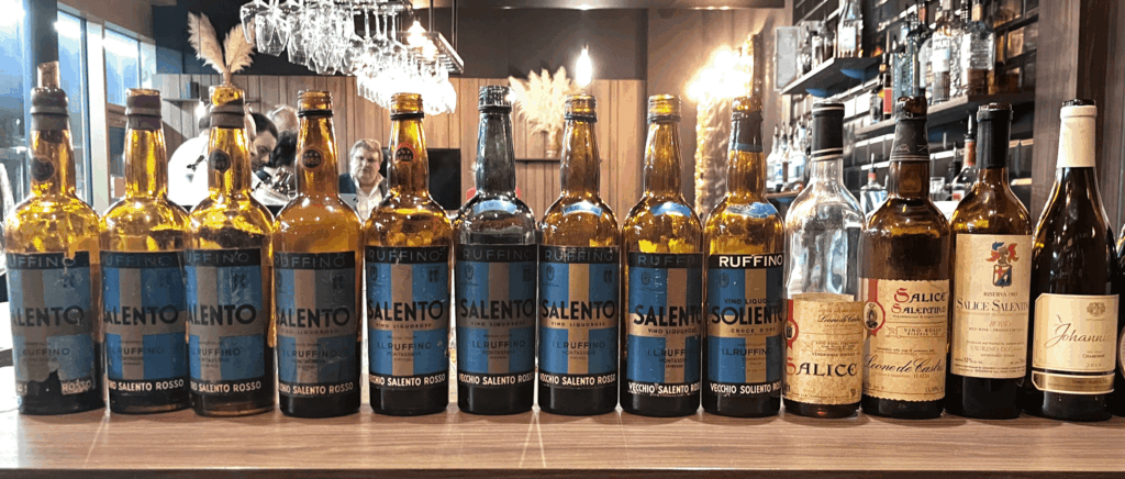 Une série de bouteilles de vins italiens des Pouilles, notamment Ruffino et Leone de Castris, alignées sur un comptoir de bar, incluant des millésimes de 1945 à 1983, présentées lors d’une dégustation à Sion