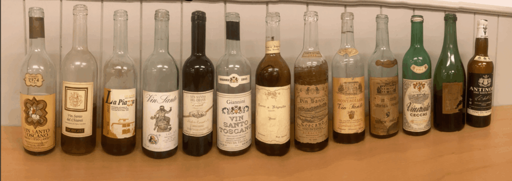 Une collection de bouteilles de Vin Santo italien, millésimes 1950 à 2011, alignées sur un comptoir en bois, avec étiquettes vieillies et bouchons en liège, devant un mur blanc à panneaux verticaux.