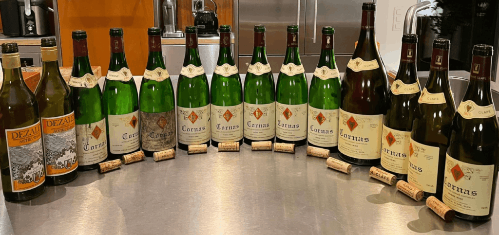 Une série verticale de bouteilles de Cornas du Domaine Clape, millésimes 1995 à 2015, alignées sur un plan de travail en acier inoxydable, avec leurs bouchons posés devant — dégustation dédiée aux collectionneurs et institutions internationales.