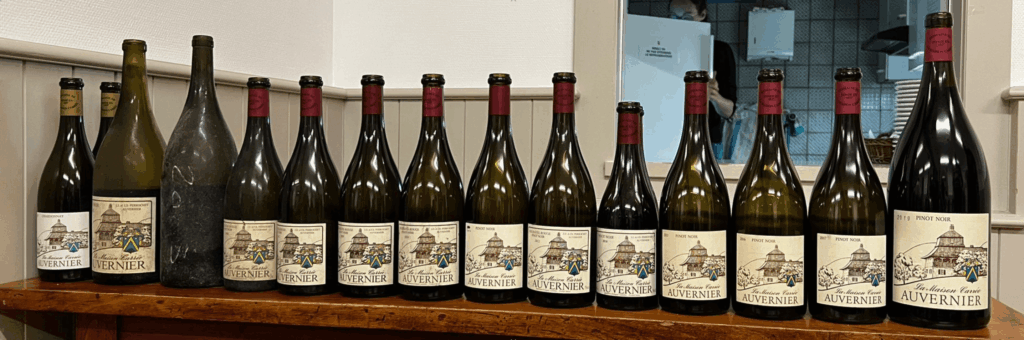 Une verticale de bouteilles de Pinot Noir « Auvernier » du Domaine La Maison Carrée, millésimes 2002 à 2019, alignées sur une étagère en bois — dégustation locale en Suisse, dédiée aux collectionneurs et amateurs passionnés du terroir neuchatelois.
