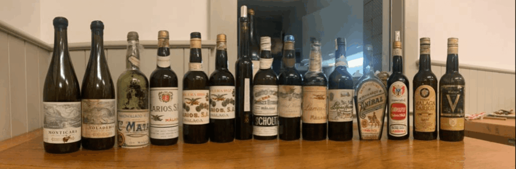 Une rangée de bouteilles de vins anciens de Malaga, présentées sur une table en bois, incluant des labels historiques comme Larios, Scholtz, Mata et Aníbal, pour une dégustation dédiée aux collectionneurs et institutions internationales.