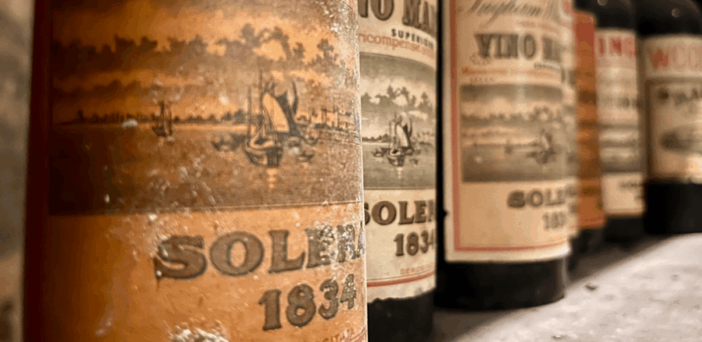 Gros plan sur une rangée de vieilles bouteilles de Marsala aux étiquettes patinées par le temps, mettant en évidence la mention "Solera 1834" et des illustrations de voiliers, témoignant de l'héritage historique de ce vin.