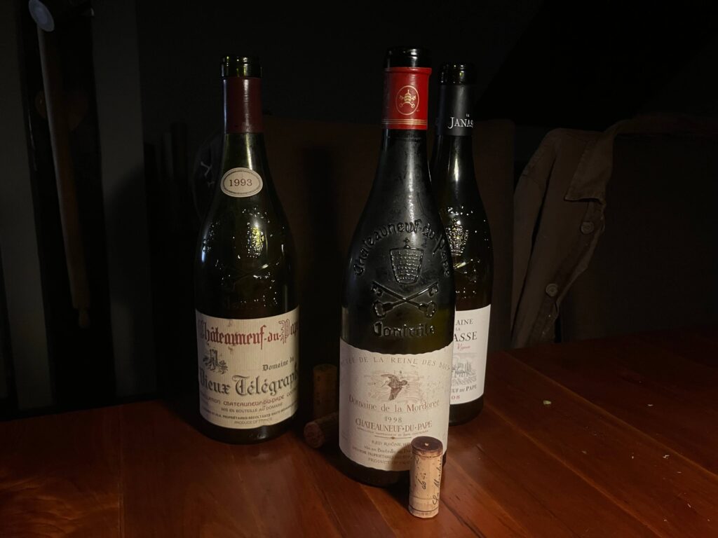 Trois bouteilles de vin rouge de l'appellation Châteauneuf-du-Pape, dont un Domaine du Vieux Télégraphe 1993 et un Domaine de la Mordorée 1998, posées sur une table en bois à côté de bouchons de liège.