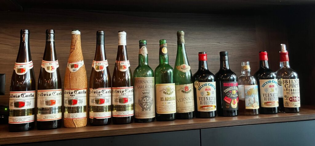 Un alignement de quatorze bouteilles de vin de Vernaccia di Oristano de la maison Silvio Carta, affichant différents millésimes et types de flacons, posées sur une étagère en bois sombre.